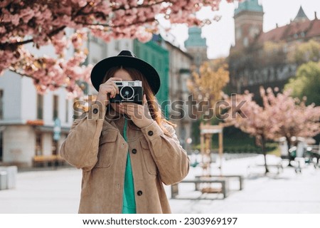 Portrait of a photographer covering her face with the camera. Carefree redhead woman with backpack posing on the street background. Positive tourist outdoors. Cheerful girl in hat is exploring city