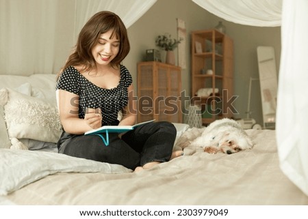 Funny picture of happy excited female making wish list in diary or writing down memories of weekend sitting on bed, while her dog feeling bored waiting for her owner to play together outside