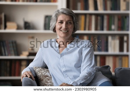 Happy attractive middle aged retired woman looking at camera, holding video call conversation with friends or family, sitting on cozy sofa at home. Relaxed mature grandmother enjoying peaceful moment. Royalty-Free Stock Photo #2303980905