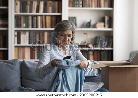 Unhappy middle aged woman unboxing carton box, looking at shirt feeling dissatisfied with getting wrong item or size, disappointed with low quality c fabric, having negative online shopping experience Royalty-Free Stock Photo #2303980917