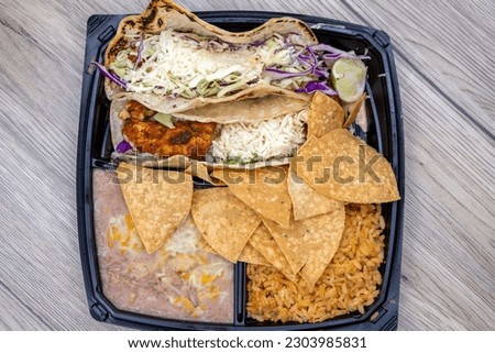Overhead view of to go order of halibut fish tacos served with rice, beans, and chips in a plastic container with dressing on the side.