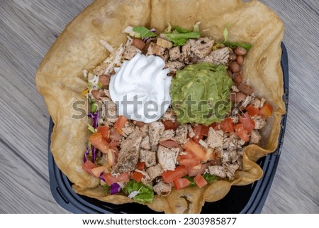 Overhead view of grilled chicken salad is served inside a crisp, tostada shell and with generous scoops of sour cream and guacamole.