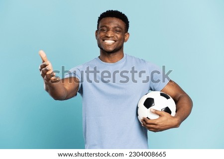 Professional african soccer player holding soccer ball, hand outstretched, gesturing isolated on blue background. Smiling male, football game concept