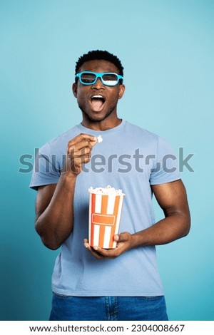 Excited nigerian man wearing 3D glasses eating popcorn watching movie isolated on blue background. Handsome hipster male in casual clothes holding paper box with snacks