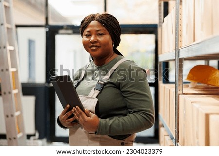 Portrait of storage room employee smiling at camera while checking customers order on tablet computer before start working at products delivery. Cheerful african american manager in storehouse Royalty-Free Stock Photo #2304023379