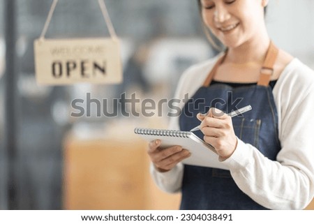 Female Asian coffee shop small business owner wearing apron standing in open sign front of counter performing stock check. afro hair employee Barista entrepreneur.
