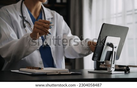 Serious female doctor using laptop and writing notes in medical journal sitting at desk. Young woman professional medic physician wearing white coat and stethoscope working on computer at workplace. Royalty-Free Stock Photo #2304058487