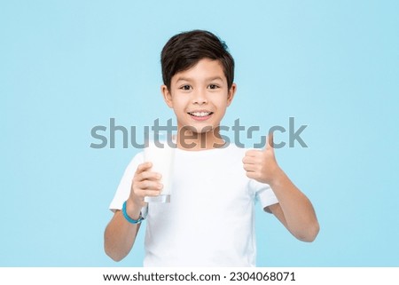 Happy smiling mixed race Asian kid boy holding a glass of fresh milk and giving thumbs up in isolated light blue color background studio shot Royalty-Free Stock Photo #2304068071