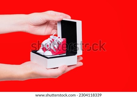 Hands holding a small gift box with a pair of tiny red high-top canvas shoes isolated on red backdrop. E-commerce and shopping related background.