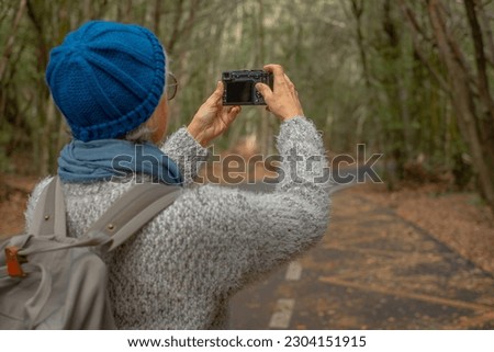 Back view of tourist photographer senior woman in outdoor excursion in the forest enjoying freedom holding a camera to take pictures