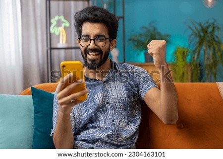 Happy indian man use mobile smartphone typing browsing say Wow yes found out great big win good news celebrate lottery jackpot doing winner gesture. Hindu young guy at home in room sitting on sofa Royalty-Free Stock Photo #2304163101