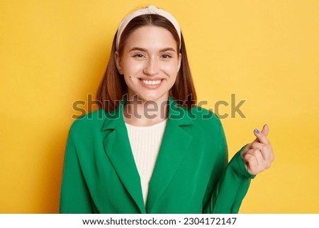 Cheerful optimistic woman wearing green jacket posing isolated over yellow background showing mini love gesture expressing feelings looking at camera with smile.