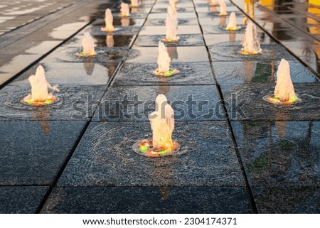 Small fountains on the sidewalk, illuminated by sunlight at sunset or sunrise in the park at summertime.