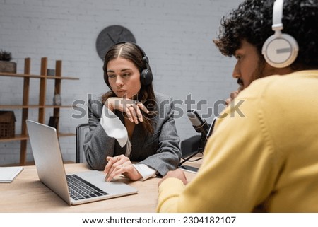 thoughtful broadcaster in headphones and grey blazer sitting near young curly indian man in yellow jumper looking at laptop near professional microphones and smartphone in modern studio