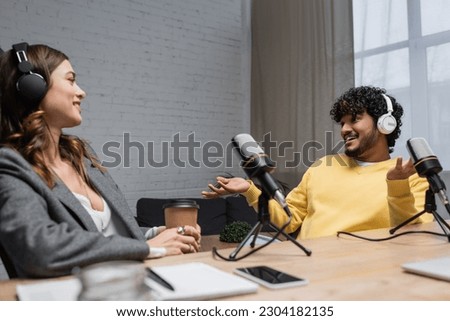joyful indian man in headphones and yellow jumper gesturing and talking to smiling colleague sitting with paper cup near microphones and blurred notebook with smartphone in broadcasting studio