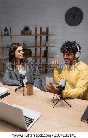 cheerful and creative indian radio host showing idea gesture while looking at mobile phone near smiling colleague and professional microphones, laptop and smartphone with blank screen in radio studio