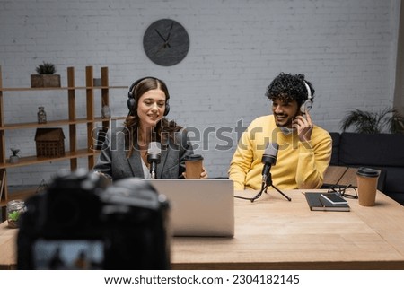 cheerful interracial podcasters in headphones working near professional microphones, notebook and mobile phone with blank screen on table with laptop and paper cups in modern studio