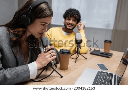 brunette radio host in headphones talking in professional microphone near laptop, smartphone with blank screen, paper cups, notebook and indian colleague smiling in studio on blurred background