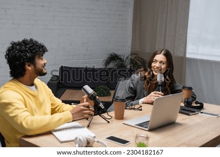 cheerful brunette radio host looking at young indian colleague while talking near microphones, laptop, notebooks, paper cups and smartphones with blank screen in studio with couch on background