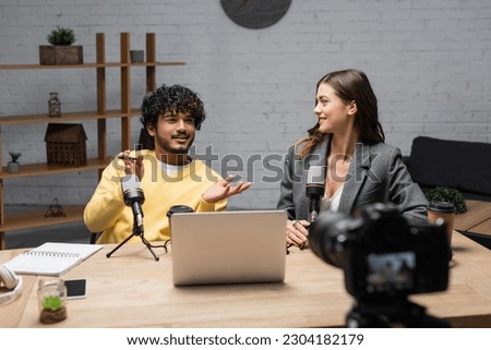 positive indian man in yellow jumper gesturing near microphones, laptop, notebook and smartphone while talking to smiling colleague sitting in blazer in front of blurred digital camera in studio