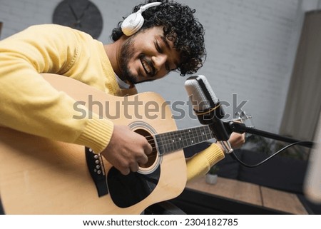 Positive and young indian podcaster in headphones playing acoustic guitar near microphone on stand during stream in podcast studio