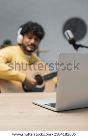focus on laptop on wooden table near young indian man in wireless headphones playing acoustic guitar while recording podcast near microphone in radio studio
