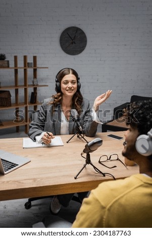 Smiling podcast host in wireless headphones talking to blurred indian guest near notebook, pen, glasses, microphones and devices with copy space during stream in studio