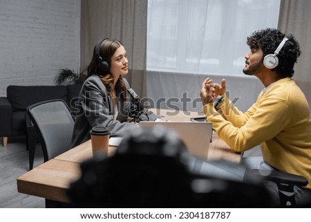 side view of smiling brunette radio host in headphones and blazer looking at curly indian man in yellow jumper talking and gesturing during interview near laptop and paper cup in modern radio studio