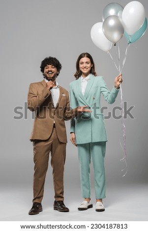 Full length of smiling young indian event host talking at microphone while standing near brunette colleague with colorful balloons on grey background