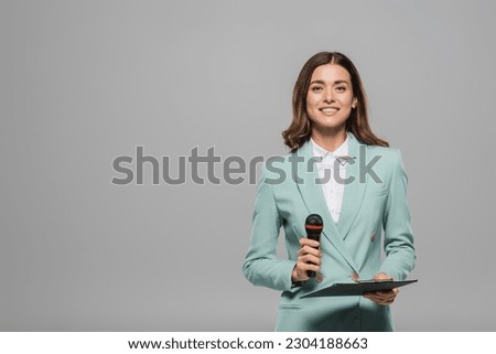Carefree and brunette event host in blue formal wear holding microphone and clipboard while looking at camera and standing isolated on grey with copy space
