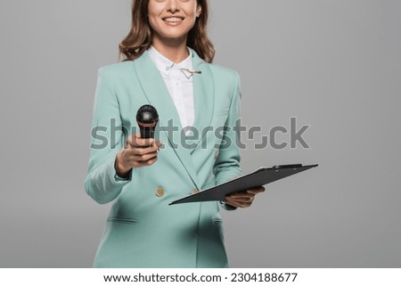 Cropped view of smiling and brunette event host in blue formal wear holding wireless microphone and clipboard during celebration isolated on grey