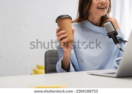 Cropped view of cheerful and brunette podcaster holding coffee to go and talking at microphone near blurred laptop on table during stream in studio