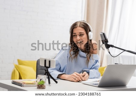 Smiling brunette podcaster in wireless headphones using smartphone on tripod and laptop near microphone and notebooks on table during stream in studio Royalty-Free Stock Photo #2304188761