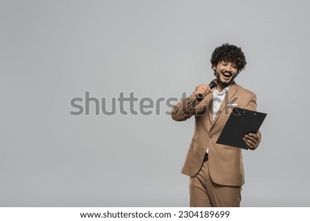 Young and overjoyed indian event host in suit talking at microphone and looking at clipboard during party while standing isolated on grey with copy space
