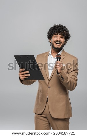 Cheerful and curly young indian host of event holding microphone and clipboard while looking at camera during celebration isolated on grey