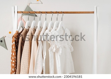 Fashion dresses and jumpers in white, beige and brown colors on hangers in wardrobe. Wooden Clothing Rack with children's outfits. Home kids wardrobe. Set of kids clothes and accessories.  Royalty-Free Stock Photo #2304196181