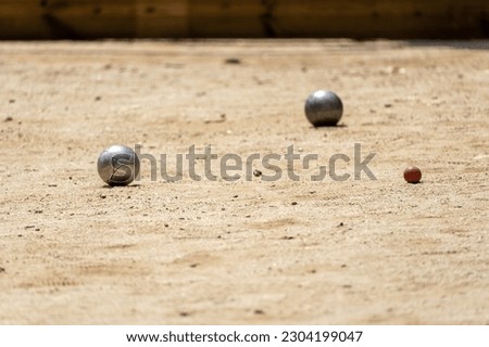 Two metal balls from the game of petanque approaching the bowling ball on a sandy ground of a petanque court on a sunny day with the shadows of the trees on the ground
