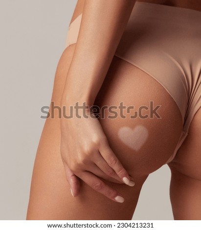 Cropped image with slim female body with tanned skin and paint heart liquid tan spray with airbrush on buttocks over white studio background. Sun protection lotion, cream. Concept of health, spf, ad Royalty-Free Stock Photo #2304213231