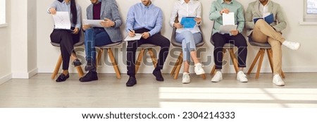 Legs of unrecognizable business people sitting on the chairs in a row with resumes or documents in their hands. Group of a staff. Job candidates seekers waiting for interview invitation turn. Banner. Royalty-Free Stock Photo #2304214333