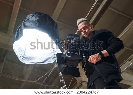 caucasian videographer holding a professional movie camera next to a lighting fixture and filming an overhead shot