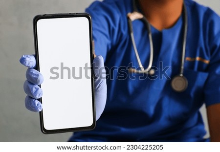 Male doctor wearing blue uniform showing smartphone with blank white screen. Medical worker demonstrating mobile phone screen, mockup