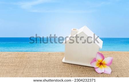 Miniature wooden house with Plumeria flower on hessian fabric over blue sea and clear sky background, tropical summer house, holiday home, summer outdoor day light, property business concept