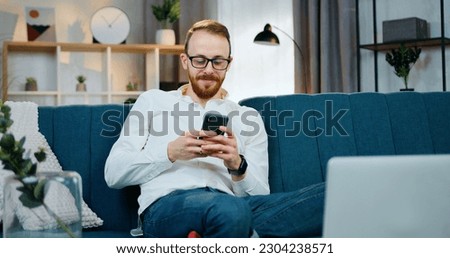 Likable smiling happy young bearded man in glasses sitting in relaxed pose on soft couch at home and browsing phone apps ,leisure concept Royalty-Free Stock Photo #2304238571