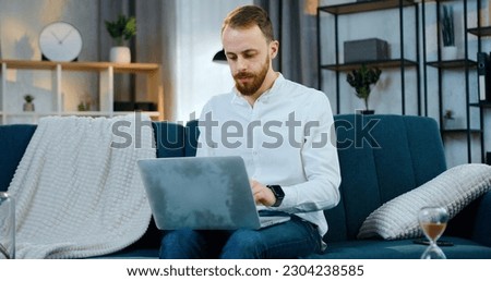 Likable serious 30-aged bearded modern man sitting on comfortable sofa at home and working with different reports and datas in computer,remote work concept Royalty-Free Stock Photo #2304238585
