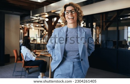 Woman standing in an office, she is smiling and wearing a suit. Business woman working with her colleagues who are having a meeting in the background.