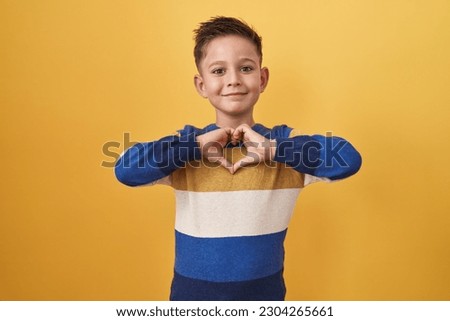 Little hispanic boy standing over yellow background smiling in love doing heart symbol shape with hands. romantic concept. 