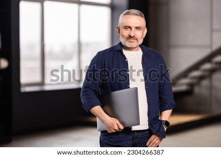 Mature man holding using laptop standing leaning on white wall, posing looking at camera. Happy adult working on pc, typing on keyboard isolated on studio background, full body length, free copy space