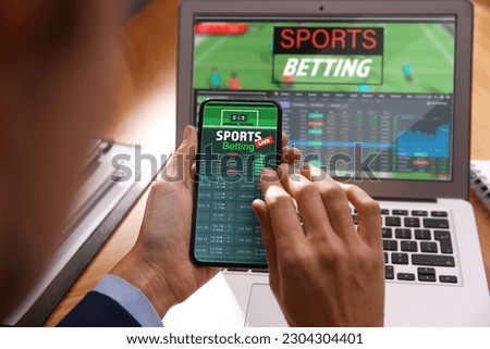 Man betting on sports using smartphone and laptop at table, closeup. Bookmaker websites on displays Royalty-Free Stock Photo #2304304401