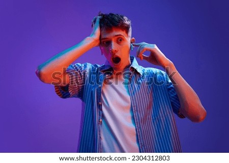 Portrait of young emotive guy in casual clothes posing in headphones with shocked facial expression against gradient purple background in neon light. Concept of human emotions, lifestyle, youth