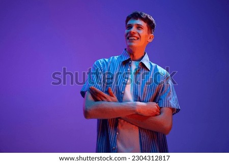Portrait of happy, smiling, young guy in casual clothes posing with positive mood against gradient purple background in neon light. Delightful. Concept of human emotions, lifestyle, youth